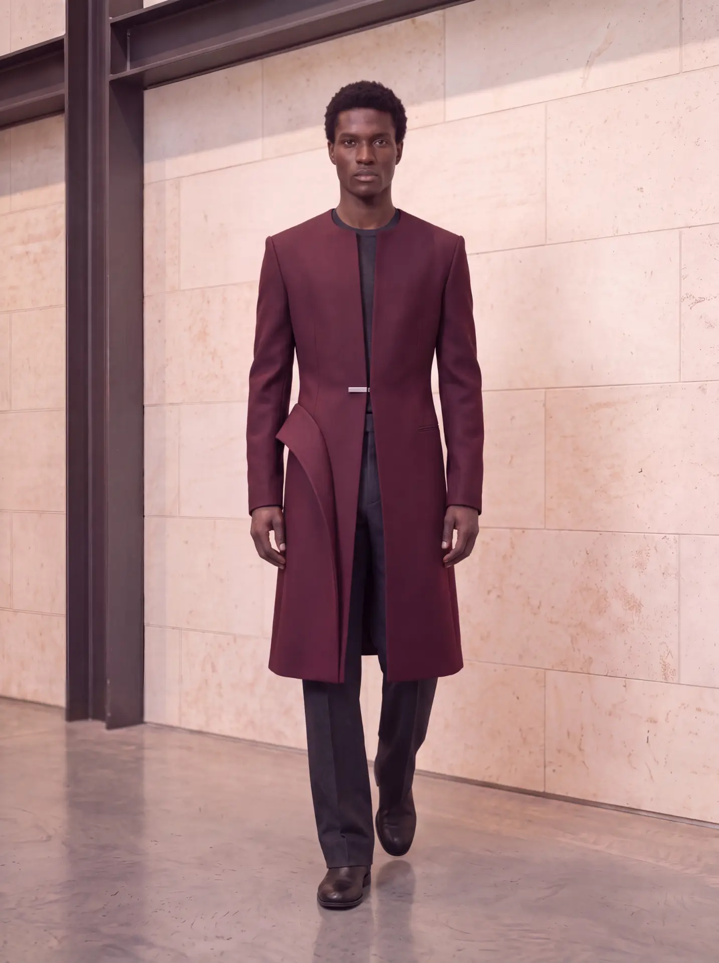 Burgundy longline coat with a sculptural hip panel and silver waist clasp over charcoal trousers